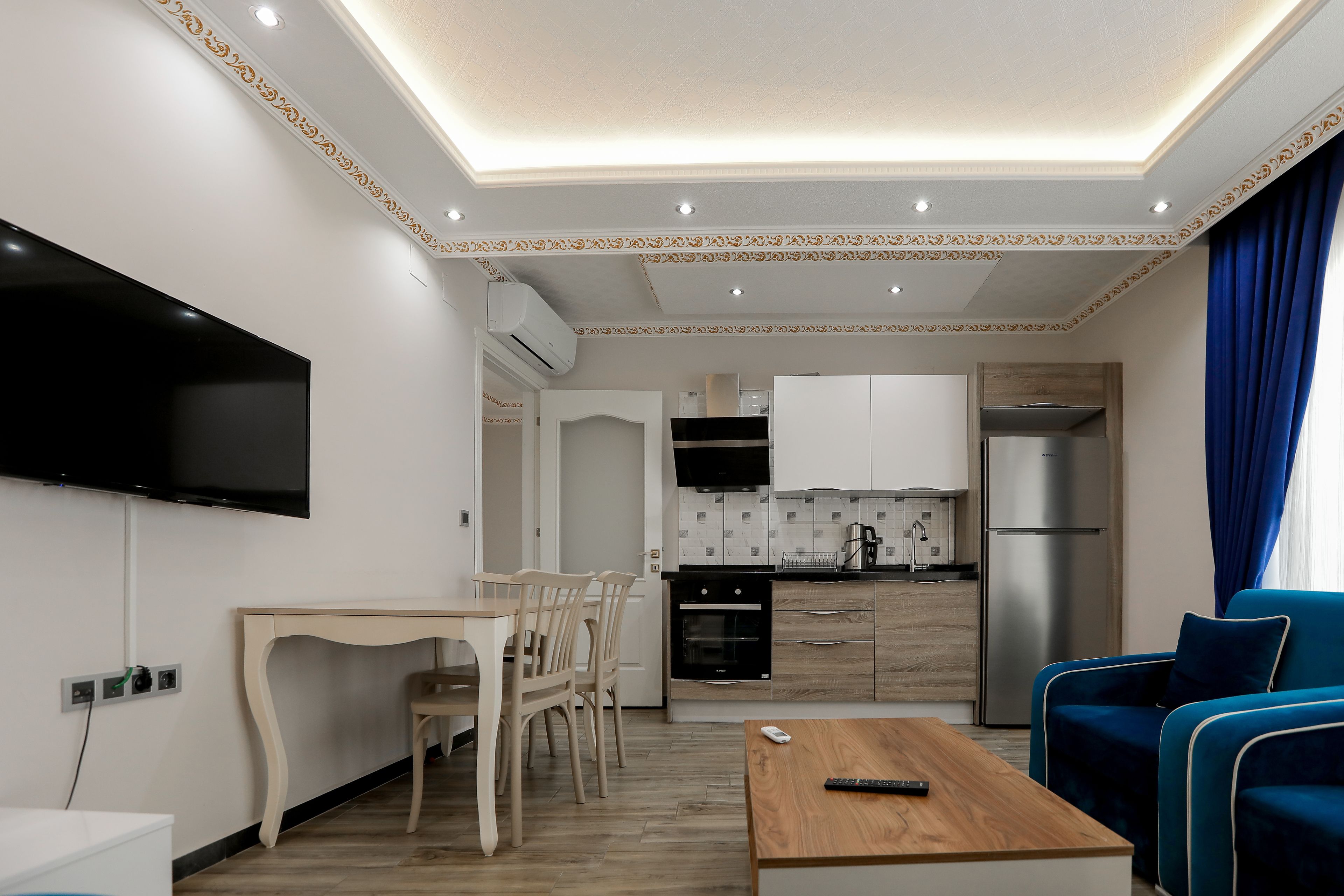 Una cocina moderna con electrodomésticos integrados, una zona de comedor con mesa y sillas de madera, y una sala de estar con un sofá azul.
