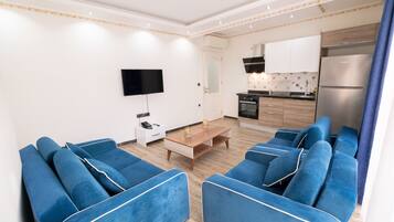 A modern living room with blue sofas, a flat-screen TV, a small table, and a kitchen area with wooden cabinets and a built-in oven.