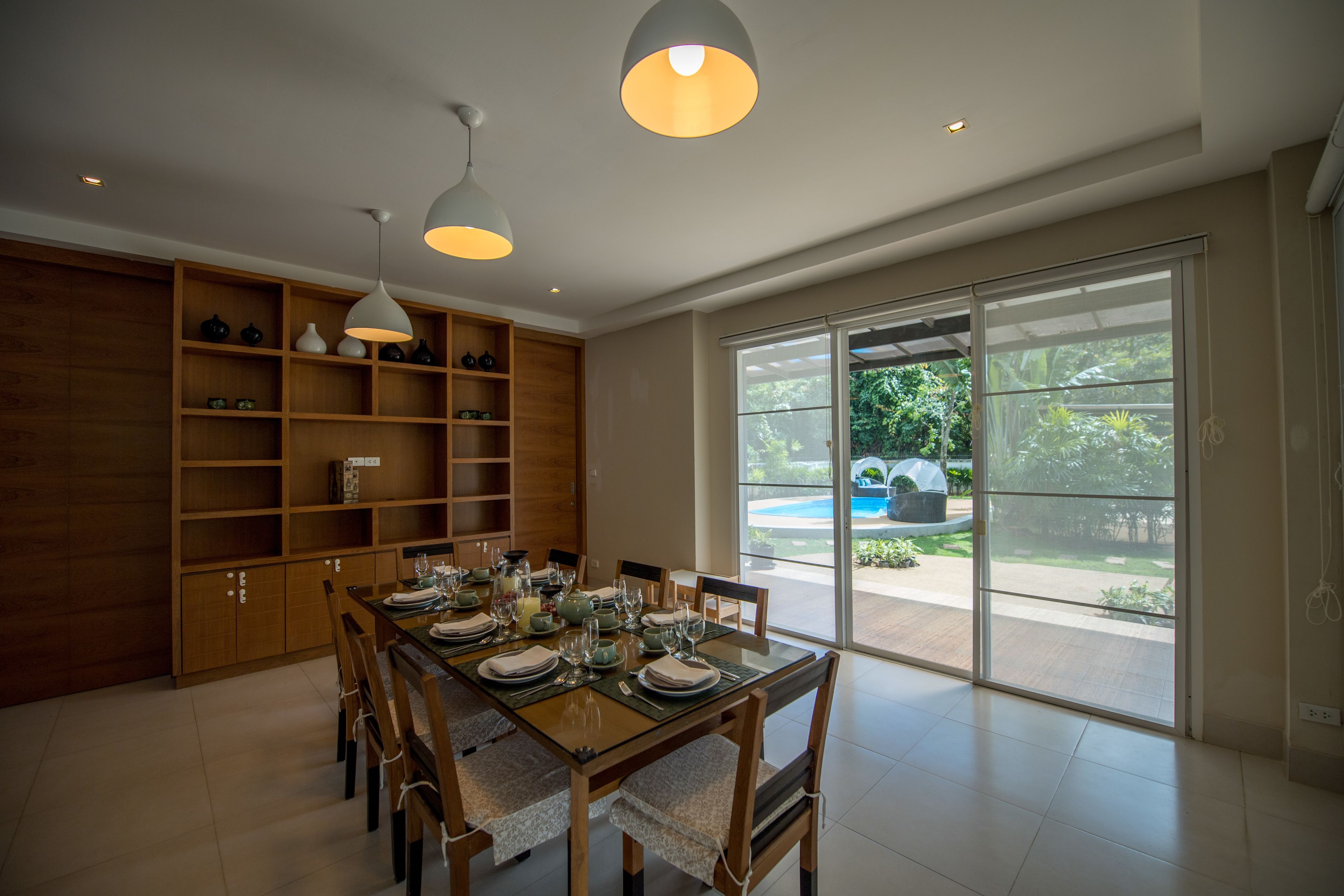 Uma área de refeições com uma mesa de madeira posta para seis pessoas, rodeada por cadeiras, e uma grande janela com vista para um jardim e piscina.