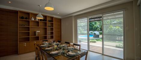 A dining area with a wooden table set for six, surrounded by chairs, and a large window offering a view of a garden and pool.