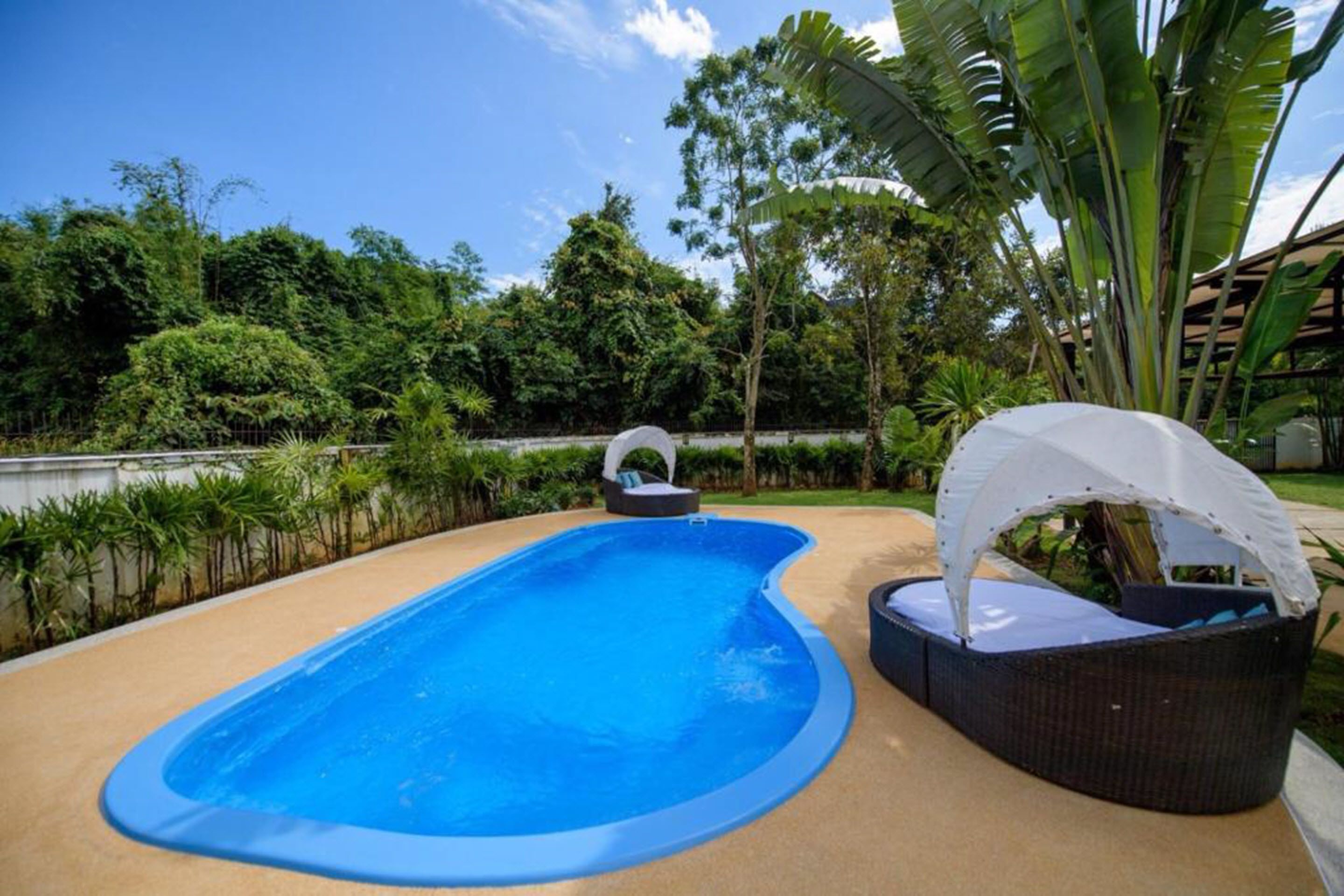 A swimming pool with a curved edge, surrounded by a beige deck. There are two lounge chairs with white canopies, one on the left and one on the right. The pool is set against a backdrop of lush greenery and trees.