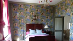 A bedroom with floral wallpaper, a wooden bed, a nightstand with a lamp, and a bathroom visible through an open door. - Château de Puichéric (Puicheric)