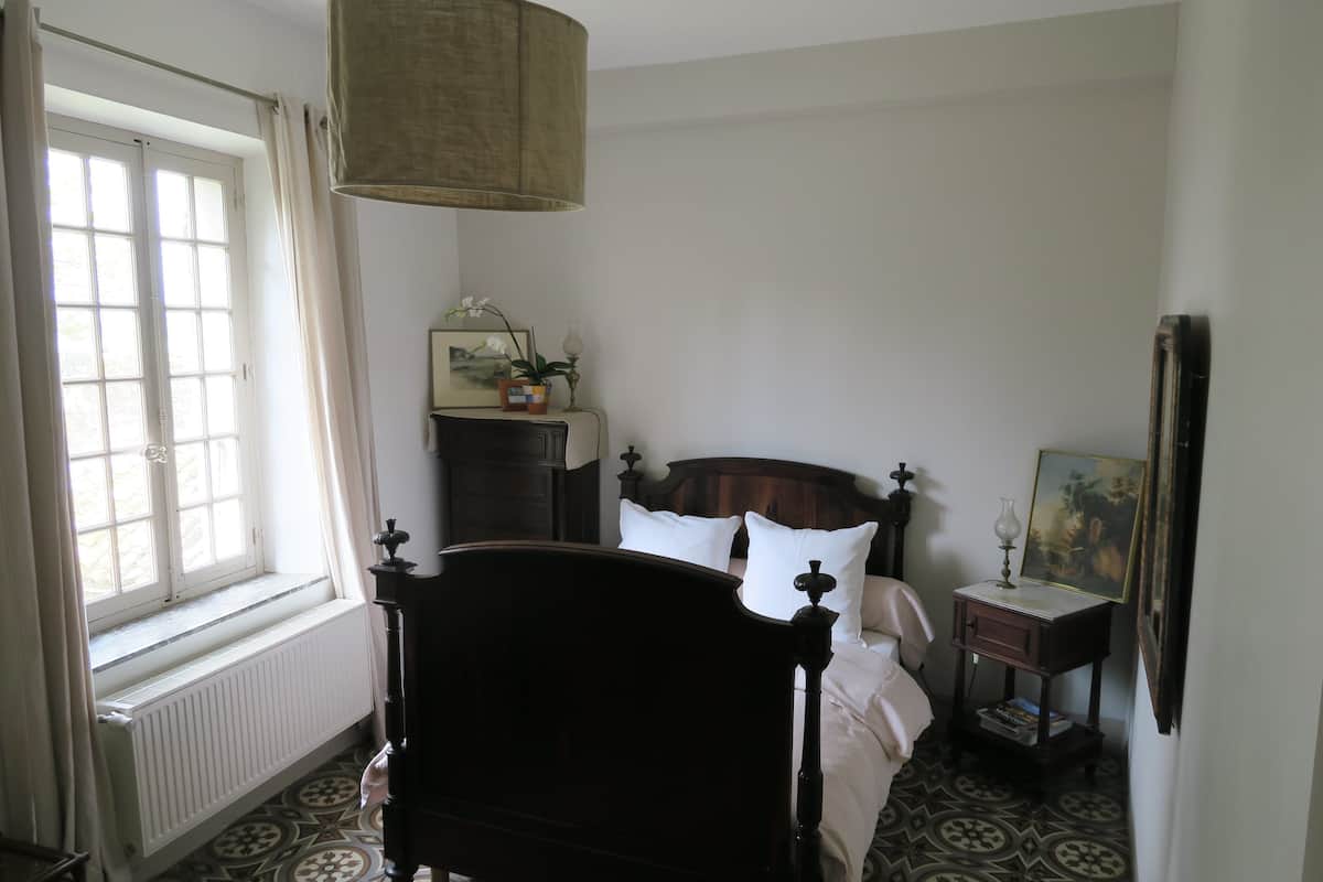 Een slaapkamer met een bed, nachtkastje, radiator, raam met gordijnen en een wandlamp.