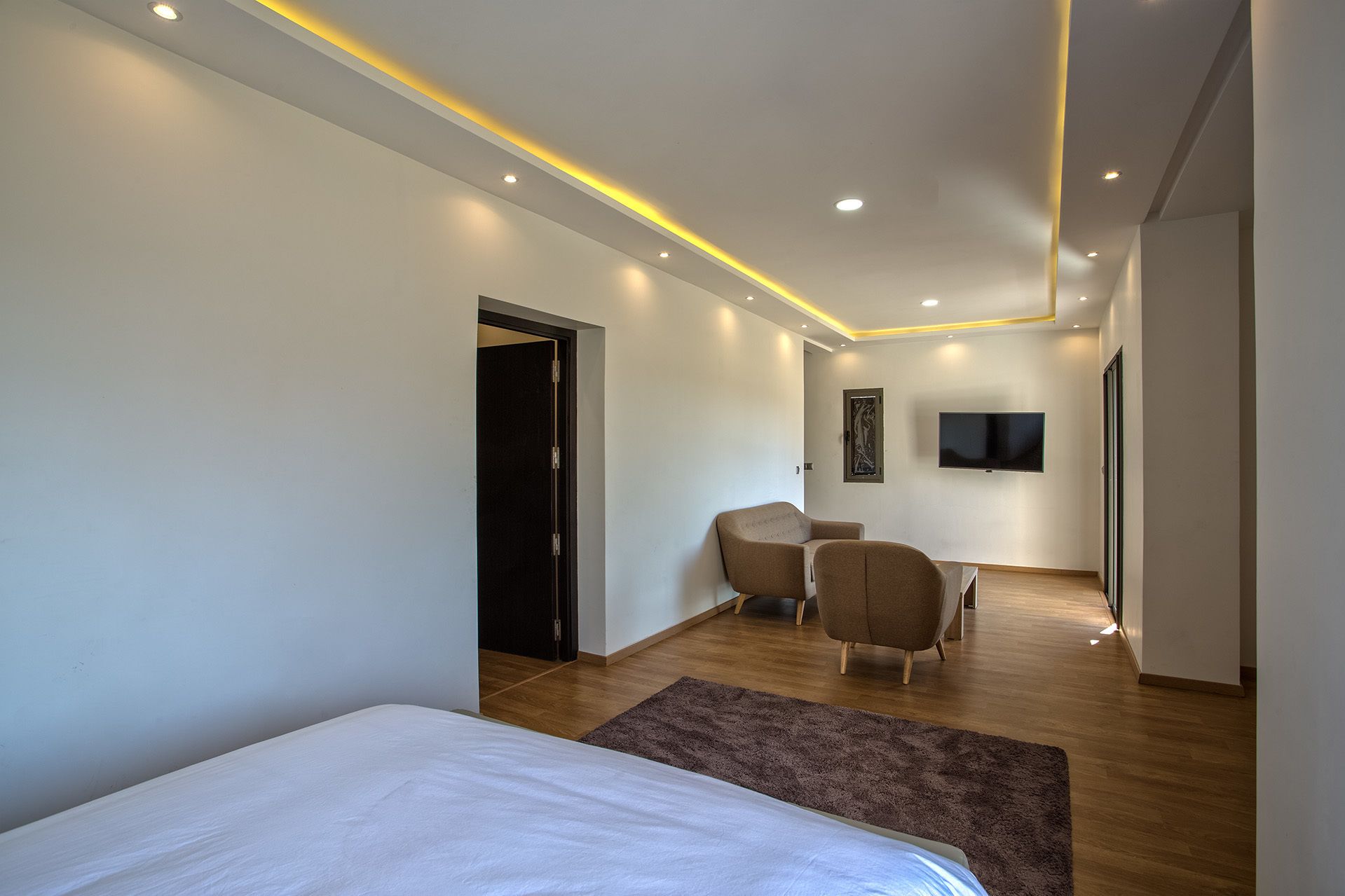 A modern hotel room with a bed, a seating area with a chair, a flat-screen TV, and a door leading to another room.