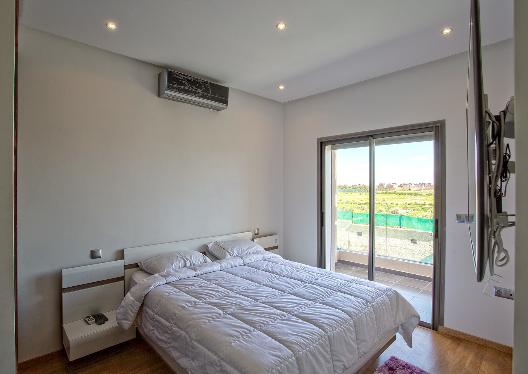 A bedroom with a bed, a bedside table, a wall-mounted air conditioner, and a sliding glass door leading to a balcony.