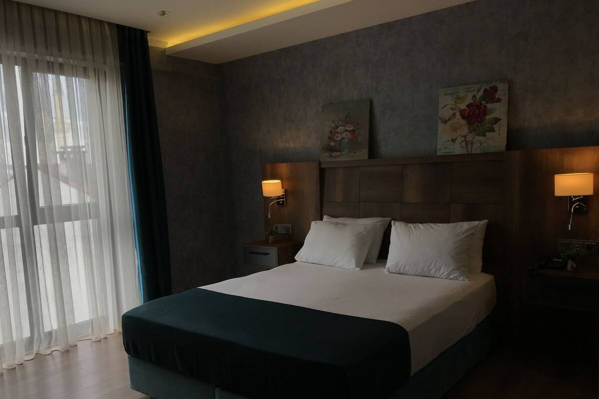 A modern hotel room with a large bed, two bedside lamps, a nightstand, and a window with sheer curtains.