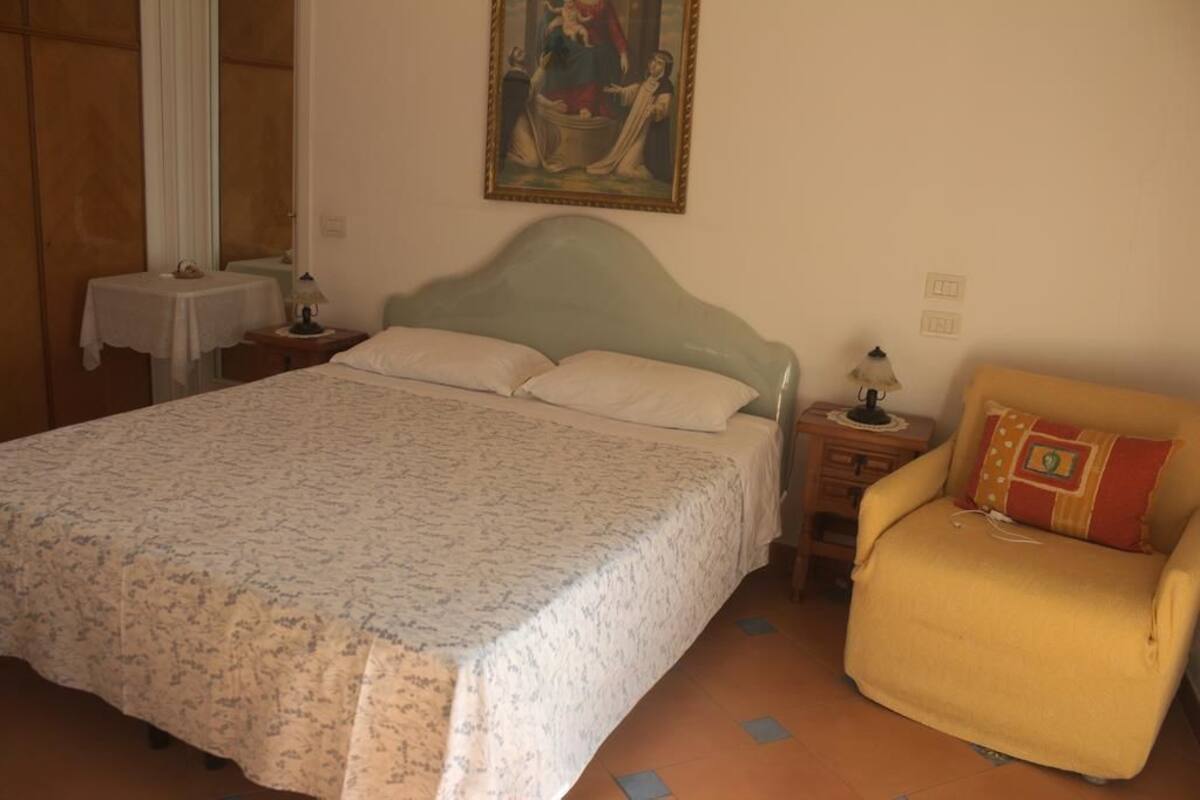 Quarto com cama, poltrona, mesa de cabeceira, luminária e um quadro na parede.