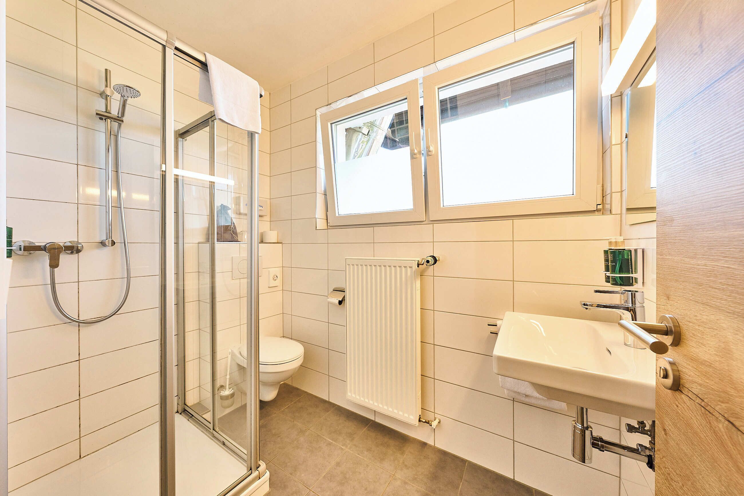 Un bagno con cabina doccia in vetro, un water, un lavandino e una finestra sopra lo specchio.