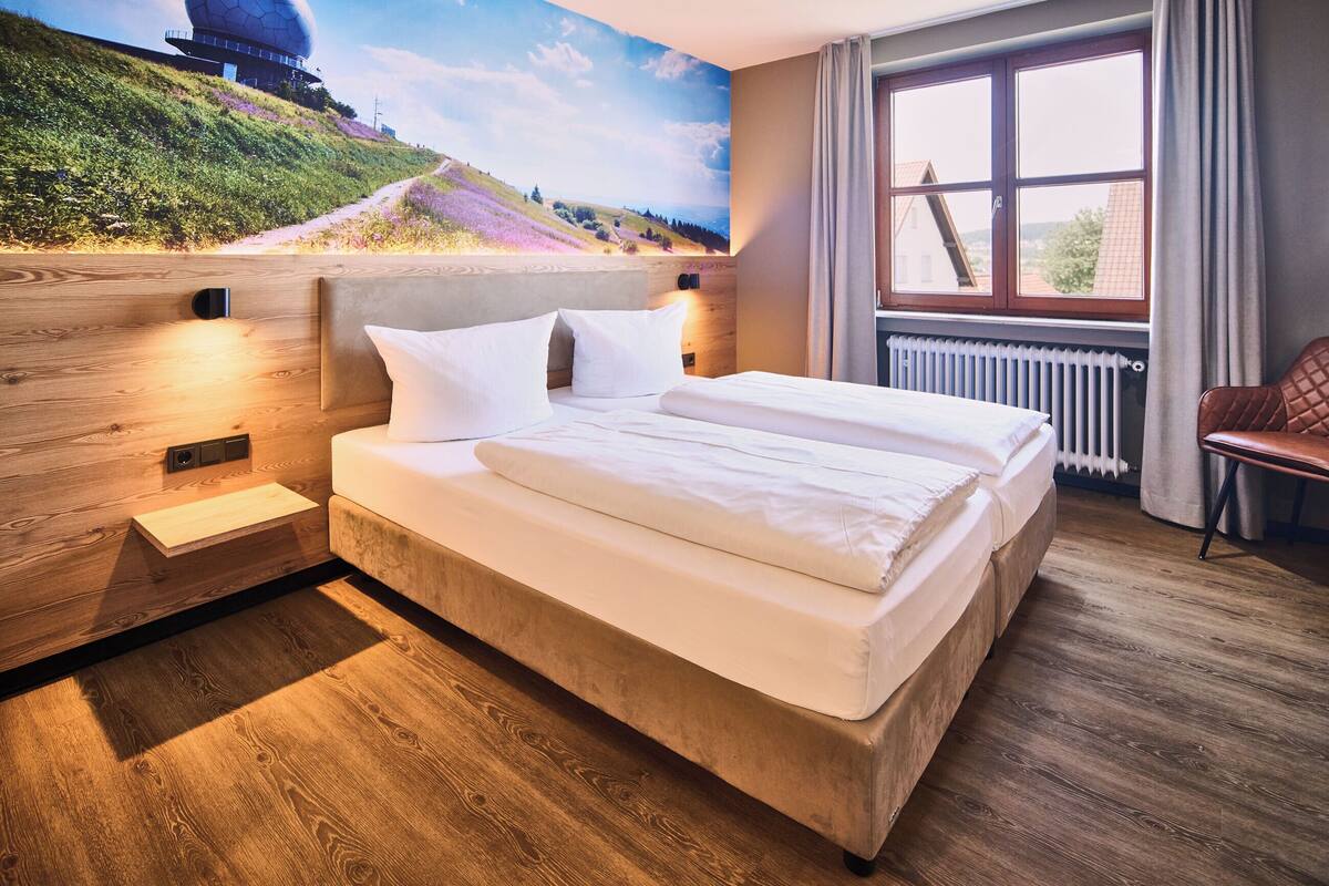 A modern hotel room with a large bed, wooden flooring, a view of a hill with a building, and a window with curtains.