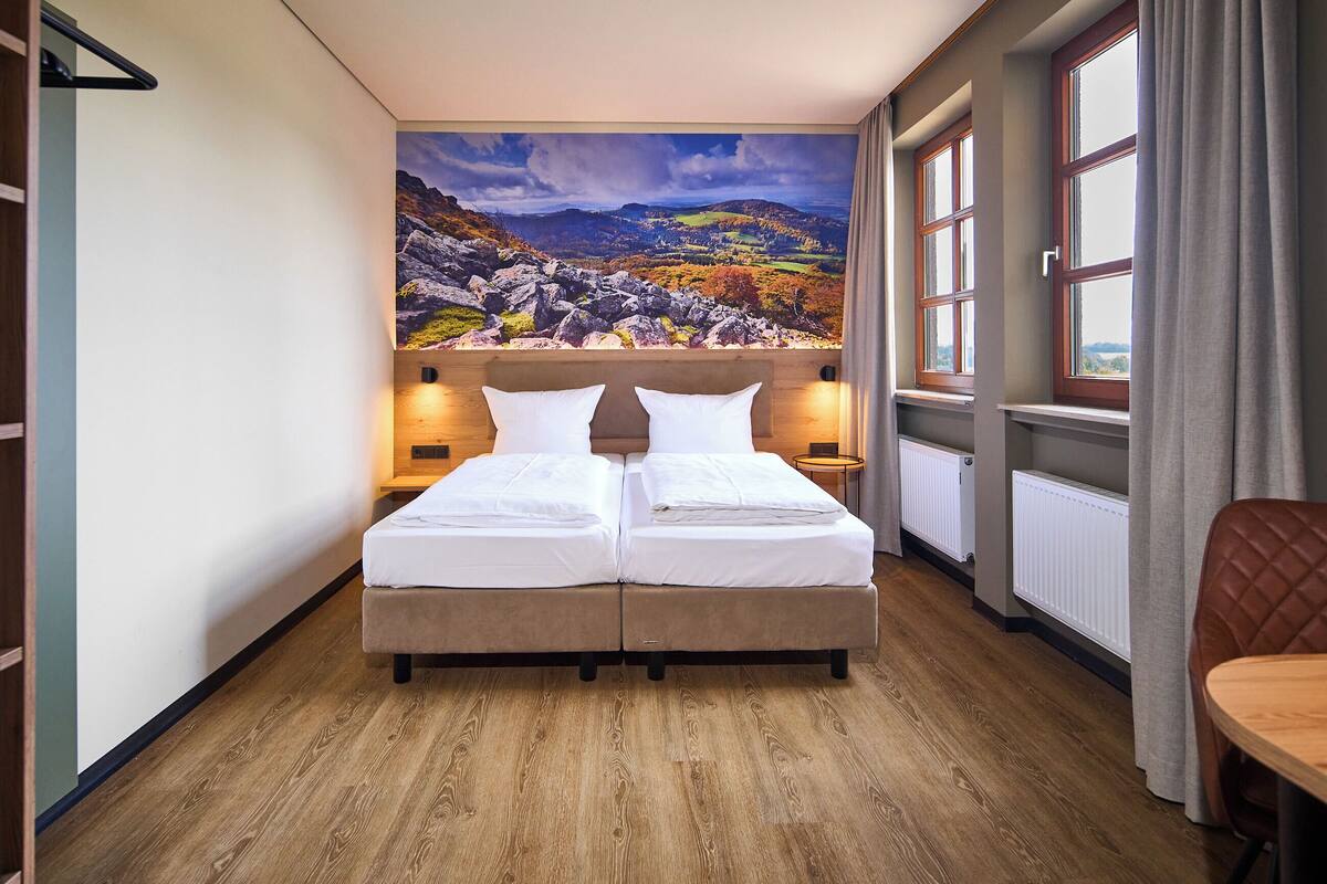 A double bed with a headboard, two bedside tables, a wooden floor, a window with curtains, and a view of a mountainous landscape.