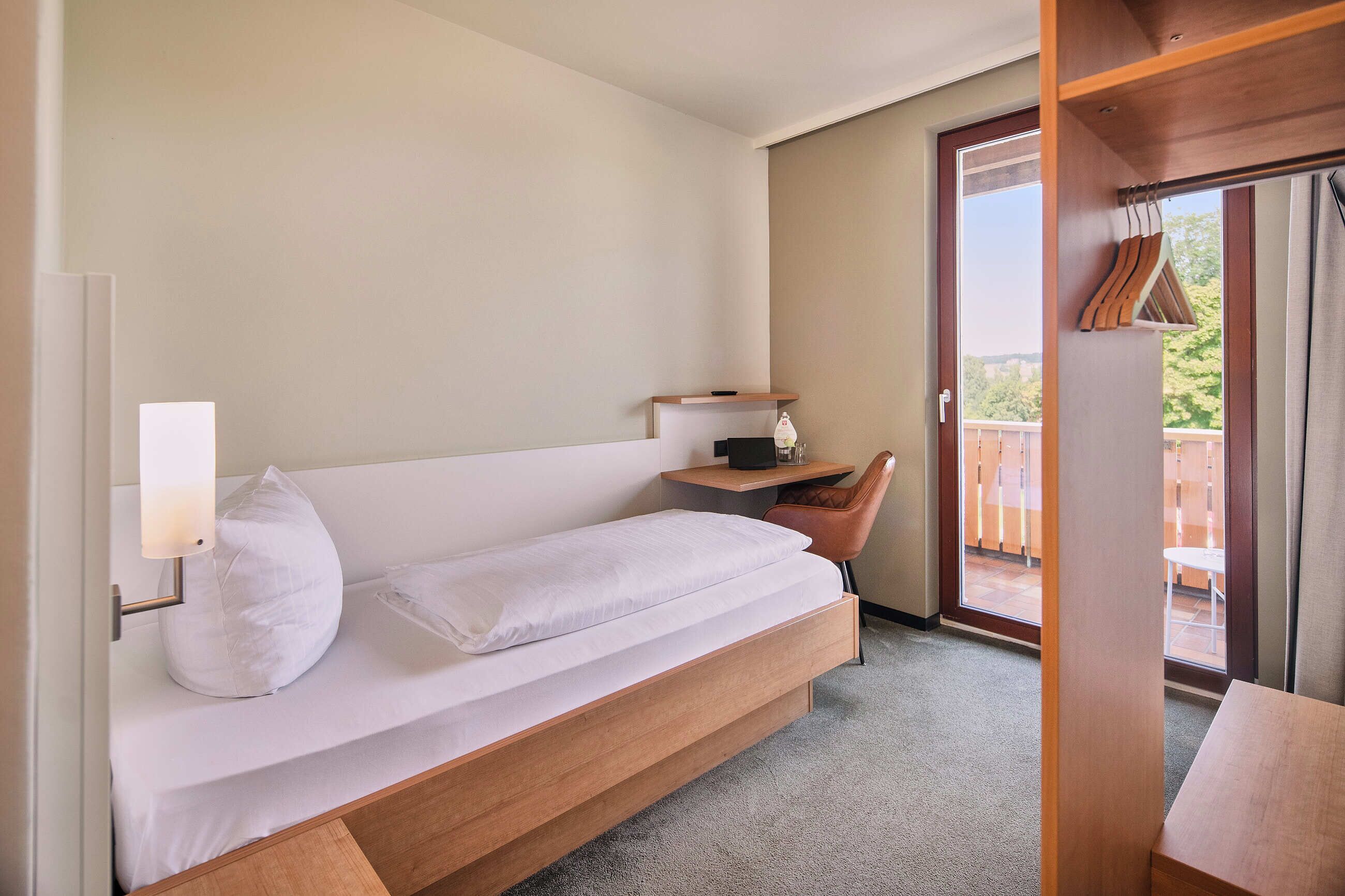 Una cama individual con estructura de madera, una mesita de noche con lámpara, y vistas a un balcón exterior.