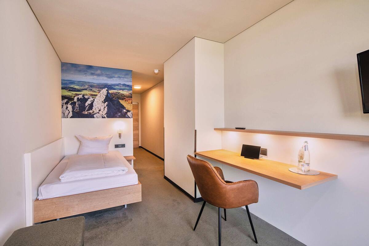 A compact hotel room with a bed, a desk, a chair, and a large landscape painting on the wall.