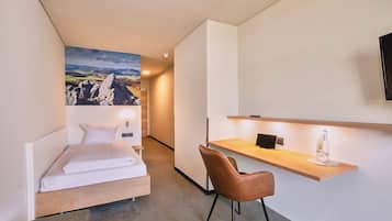 A compact hotel room with a bed, a desk, a chair, and a large landscape painting on the wall.