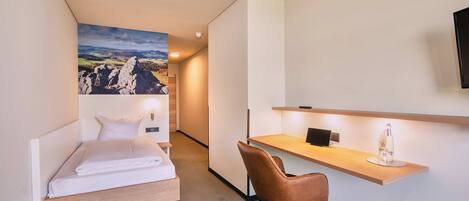 Una habitación de hotel compacta con una cama, un escritorio, una silla y un cuadro grande de paisaje en la pared.
