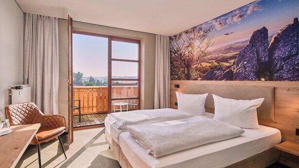 A hotel room with a large bed, a wooden headboard, a desk with a chair, and a view of the outdoors through a sliding glass door. - Hotel-Restaurant Berghof (Petersberg)