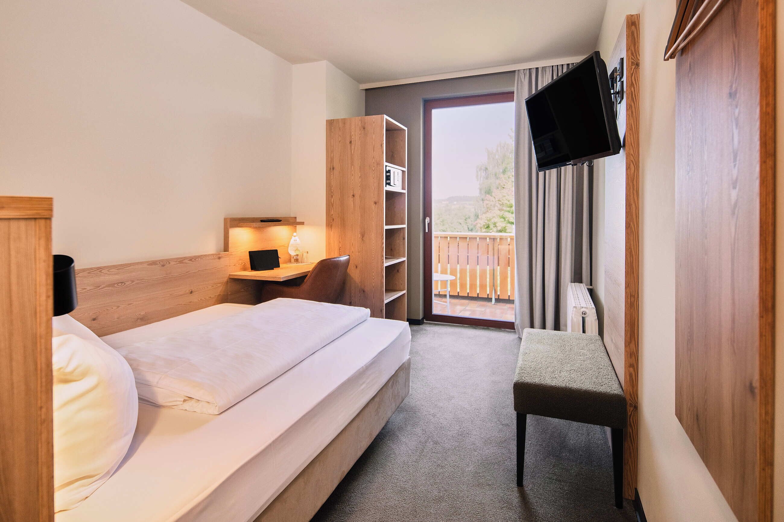 Habitación de hotel con cama, escritorio, silla, televisor montado en la pared y balcón con vistas.