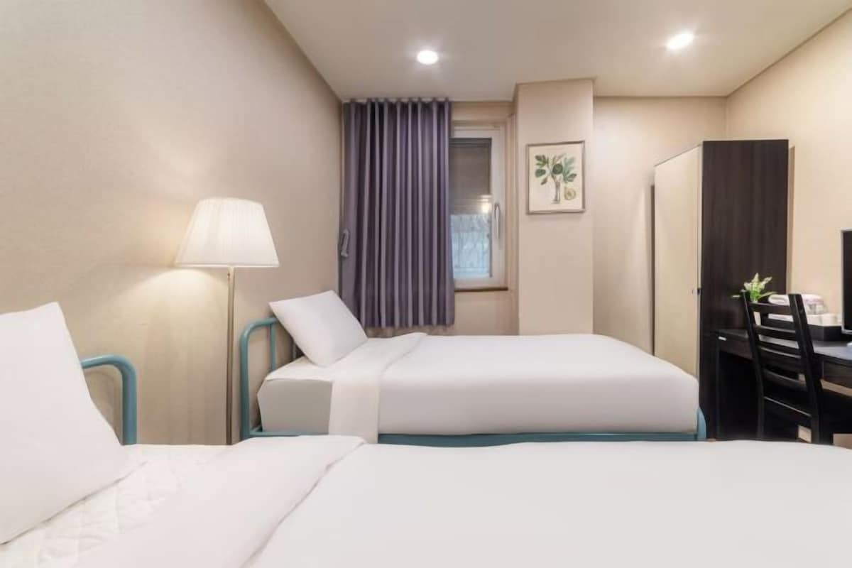 Habitación de hotel con dos camas, un escritorio, una silla, una lámpara, una ventana con cortinas y un cuadro en la pared.