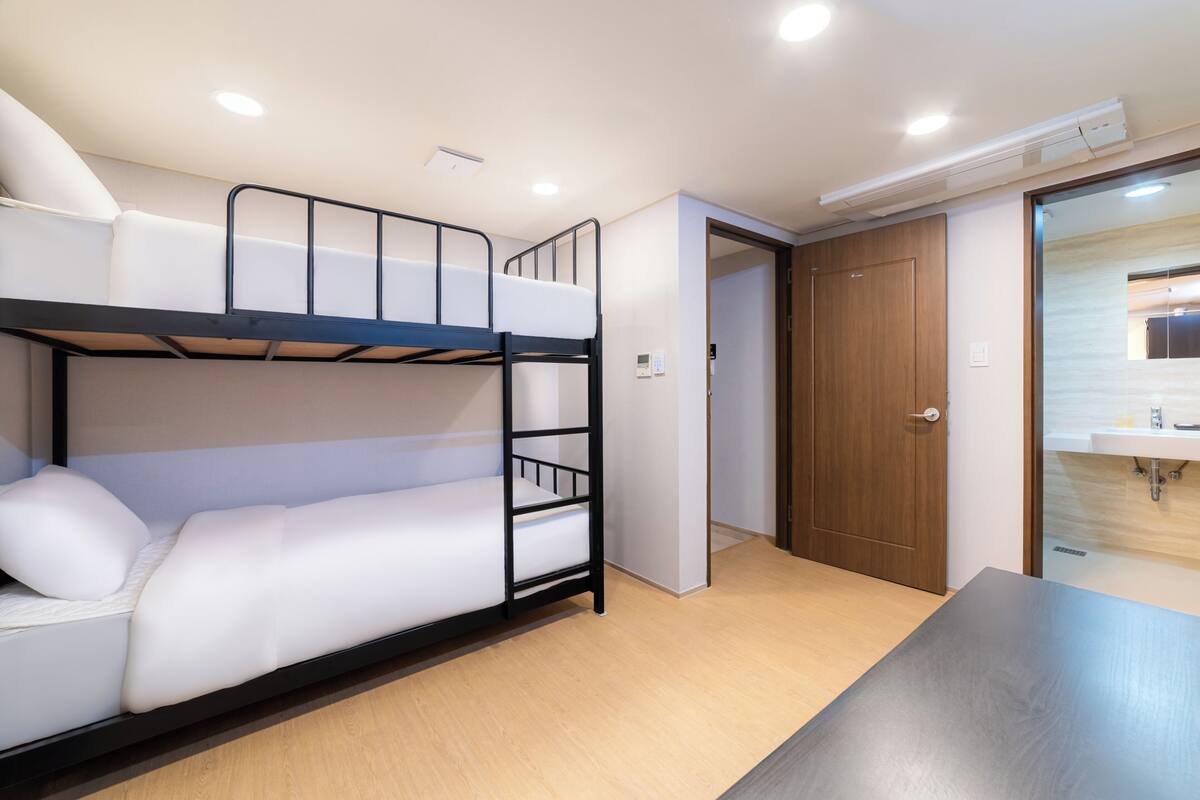 Une chambre avec des lits superposés, une porte en bois, une petite table et une salle de bain visible à travers une porte en verre.
