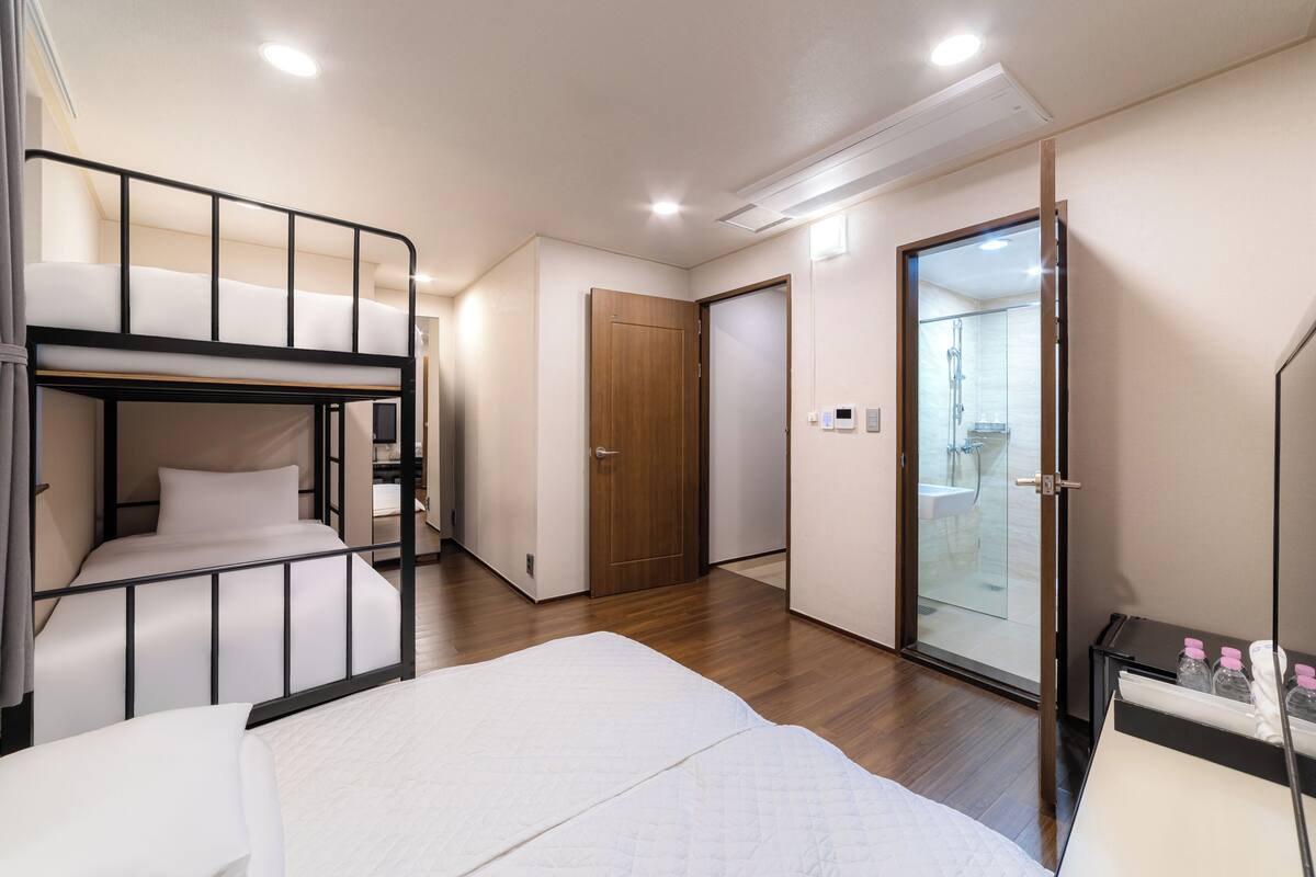 Una habitación con una cama litera, una cama individual, una puerta de madera y un baño con ducha de vidrio.