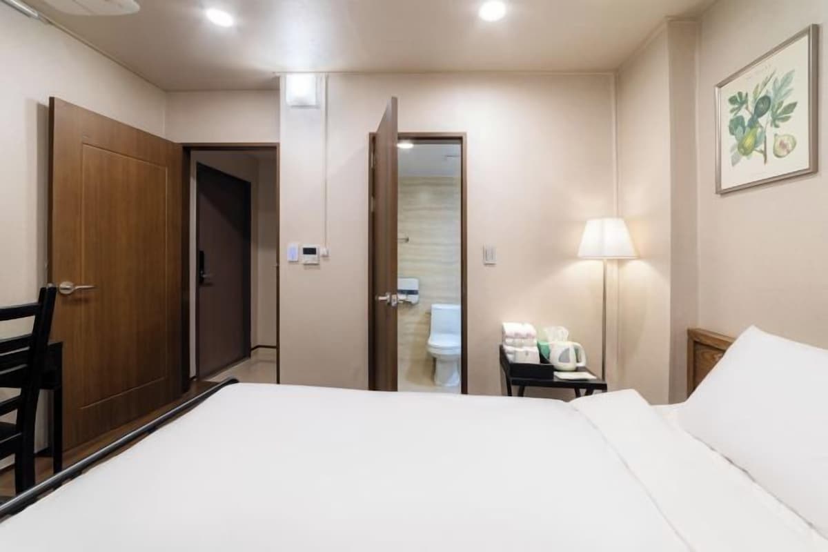 Habitación de hotel con cama, escritorio con lámpara, silla, puerta de madera y baño visible a través de un acceso abierto.