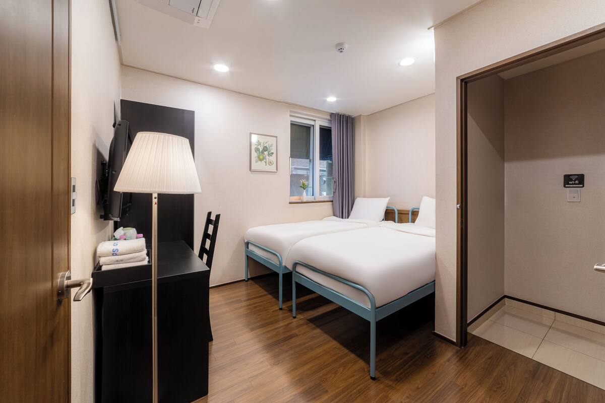 Habitación de hotel con dos camas, un escritorio con lámpara, una ventana con cortinas y una puerta con el letrero de Wi-Fi.