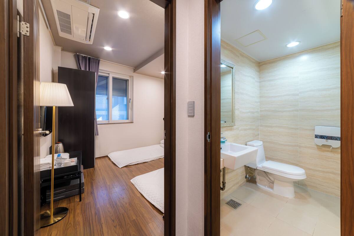 Habitación de hotel con cama, mesita de noche, lámpara, ventana y baño con lavamanos e inodoro.