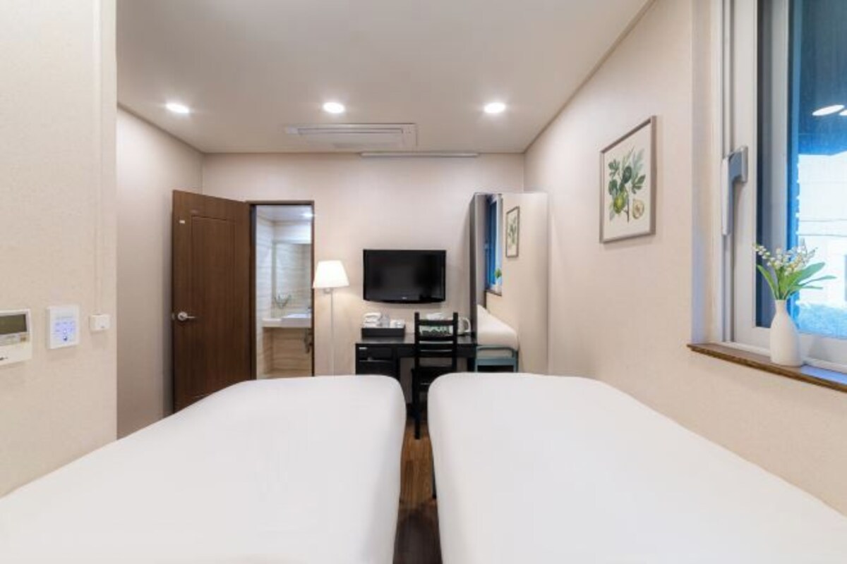 A hotel room with two beds, a TV, a desk, a lamp, and a window with a plant.