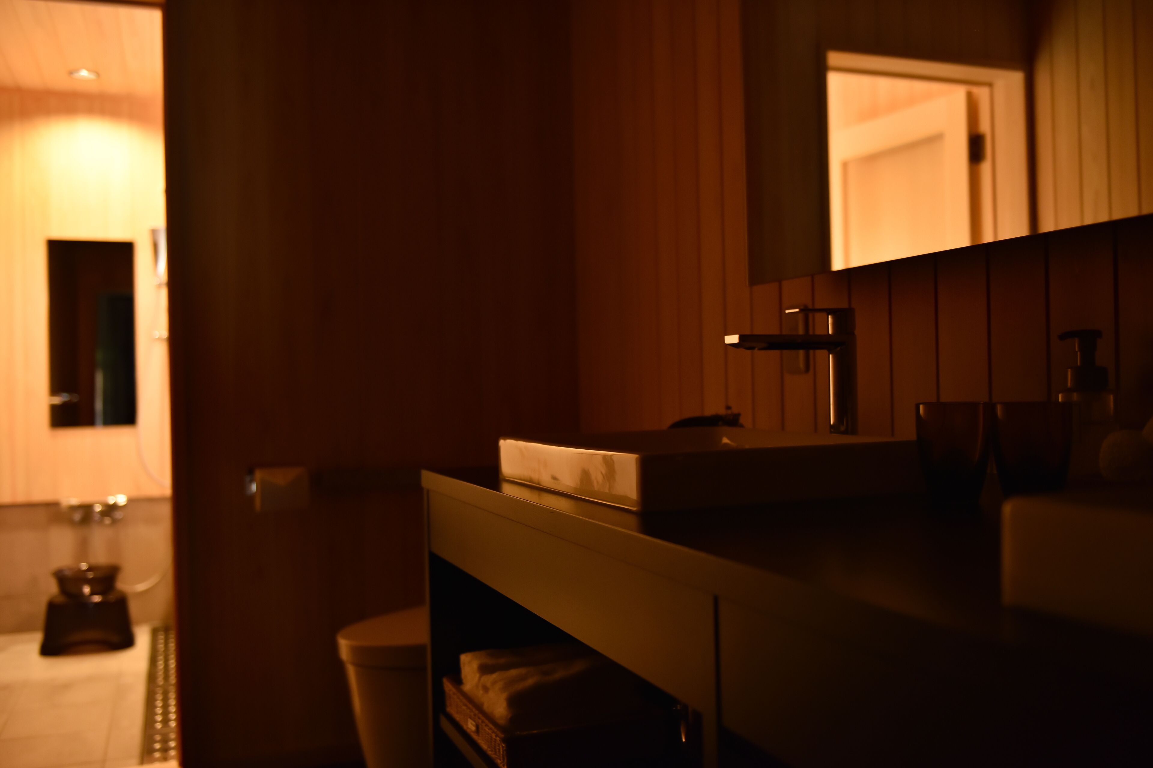 A dimly lit room with a wooden floor, a white bench, and a small table with a bottle and a tissue box.