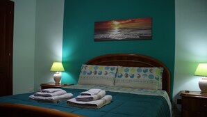 Een netjes opgemaakt bed met een blauwgroene deken en kussens, twee bedlampjes en een schilderij van een zonsondergang boven de oceaan aan de muur.