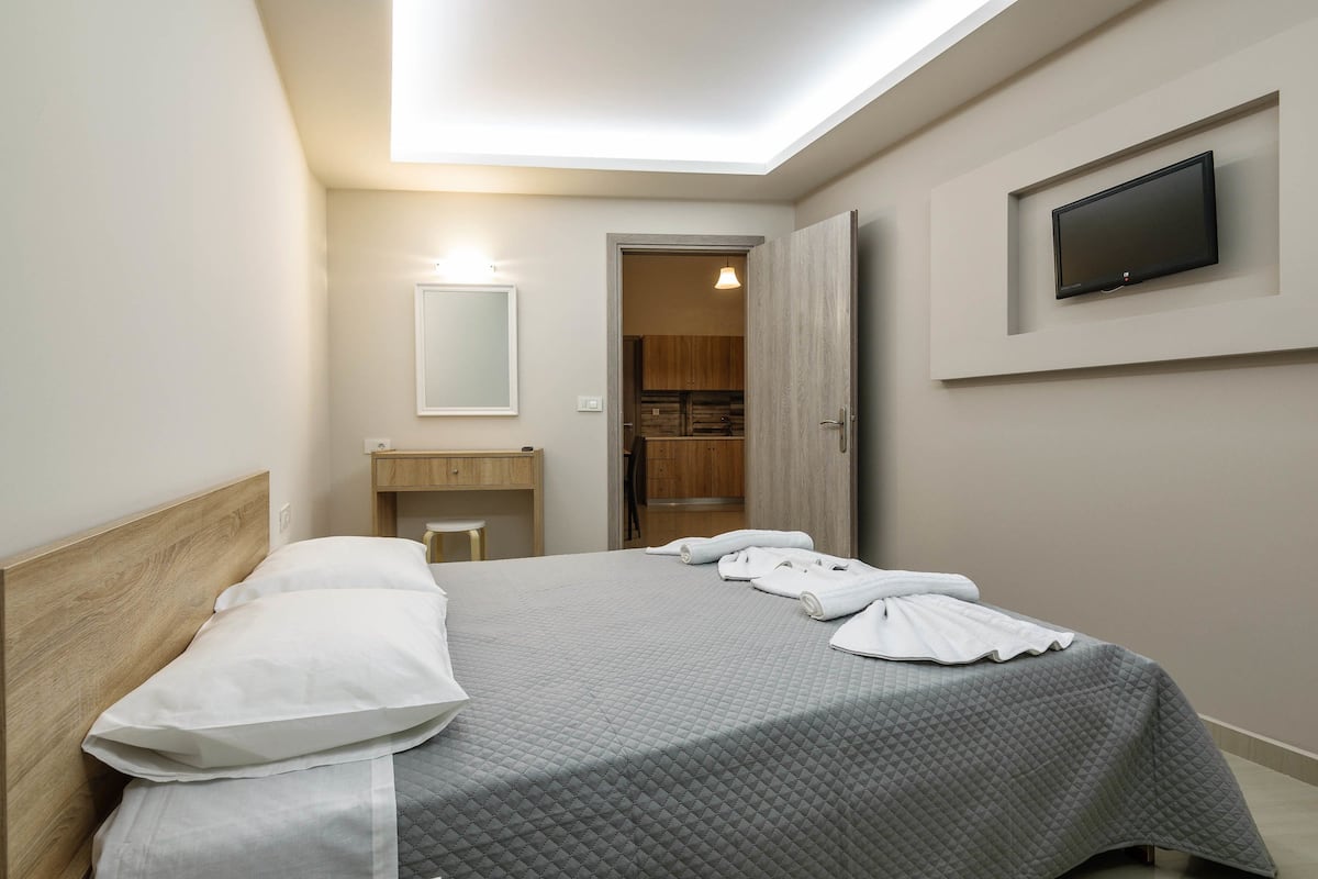 A neatly made bed with a gray quilt, a wooden headboard, and white pillows. A flat-screen TV is mounted on the wall above the bed.