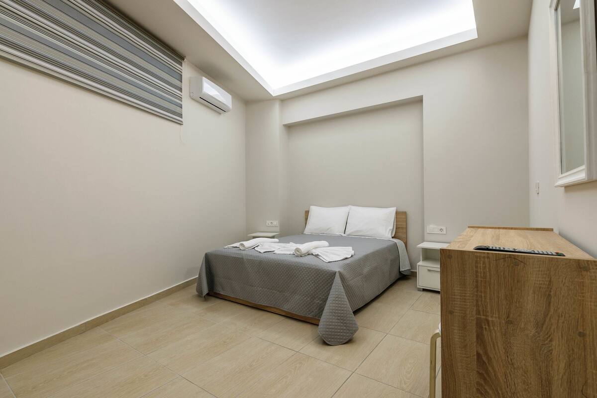 A bedroom with a skylight, a bed with a grey bedspread, a wooden desk, and a nightstand.