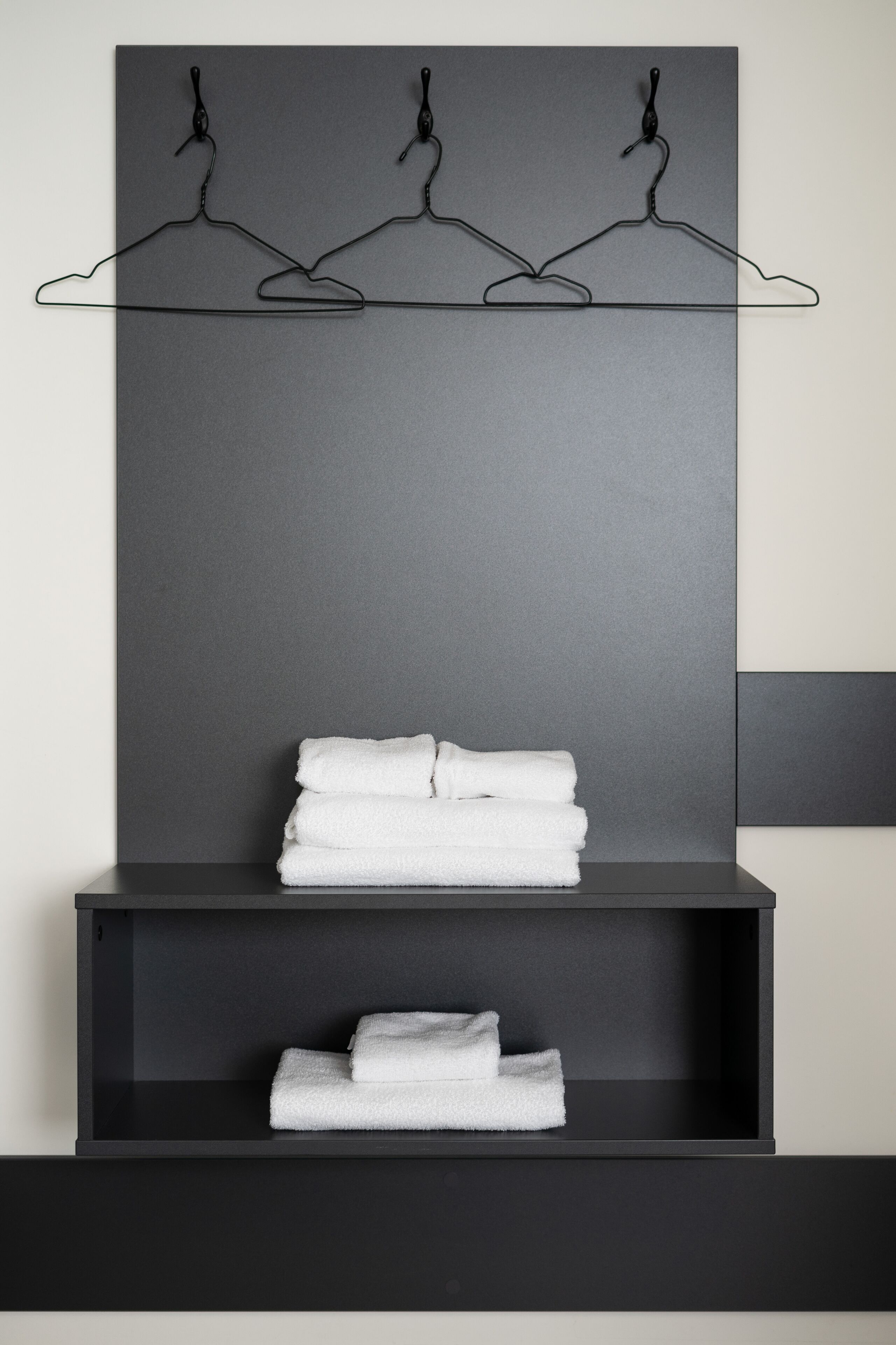 A modern bathroom with a dark gray wall, a black shelf with folded white towels, and three black wall-mounted towel hooks.