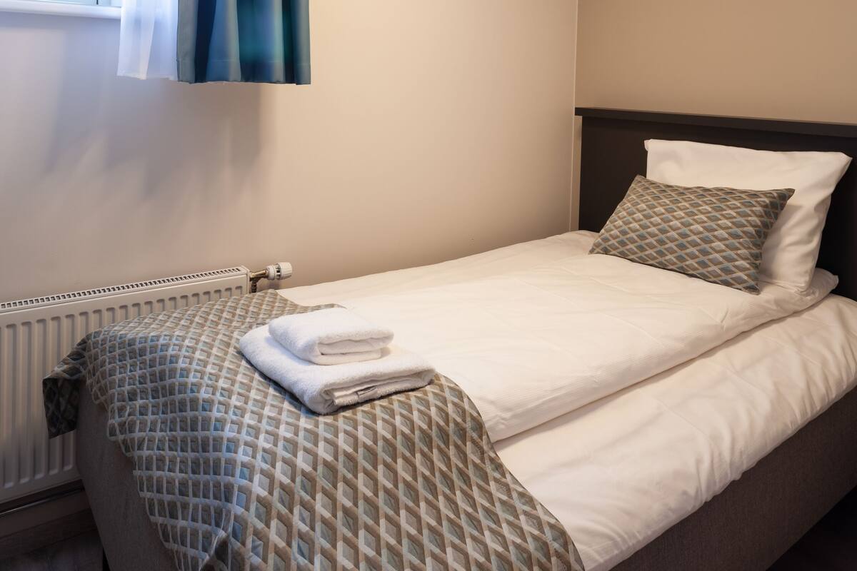 A neatly made bed with a patterned bedspread, a pillow, and a folded towel. There is a window with blue curtains, a radiator, and a ceiling light fixture.