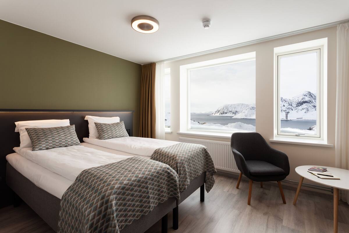 A hotel room with two beds, a chair, a small table, and large windows with a snowy mountain view.