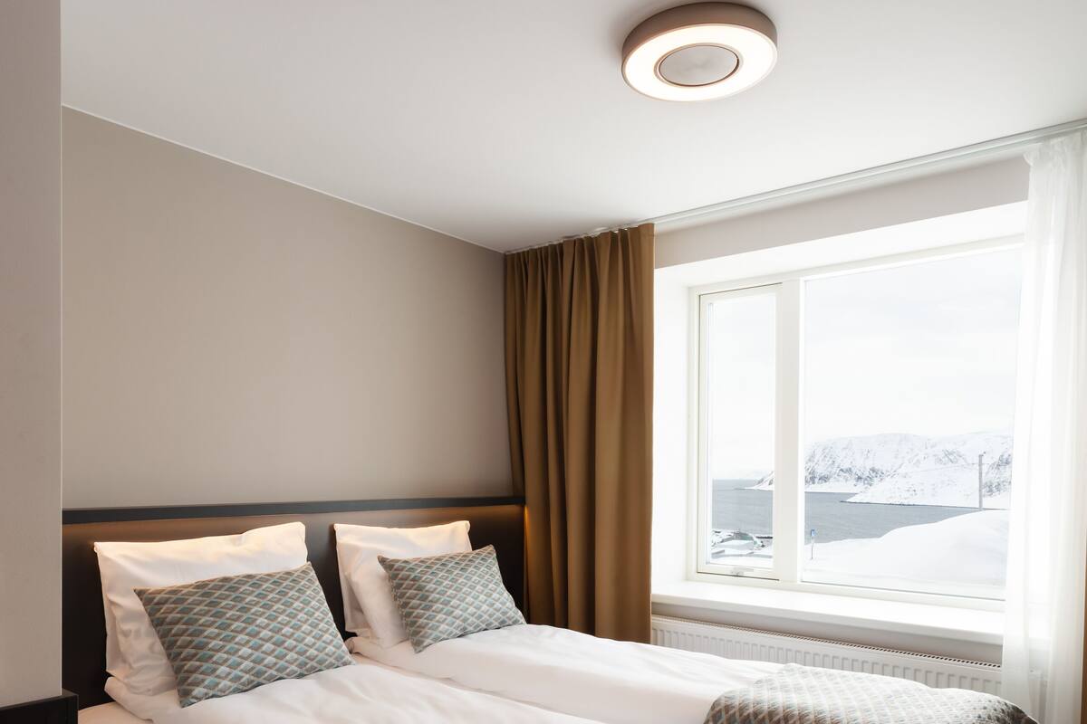 A hotel room with a large bed, a window with a view of a snowy landscape, and a ceiling light.