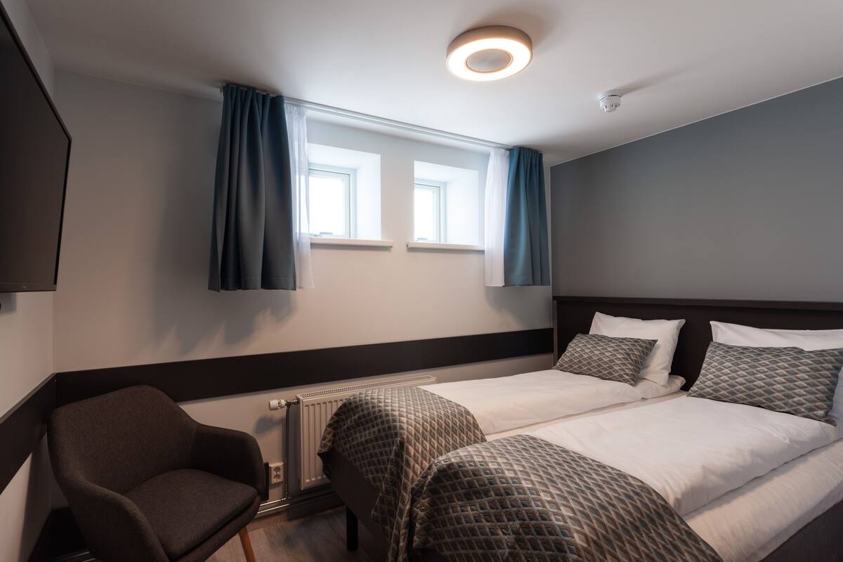 A hotel room with two beds, a chair, a radiator, and a window with curtains.