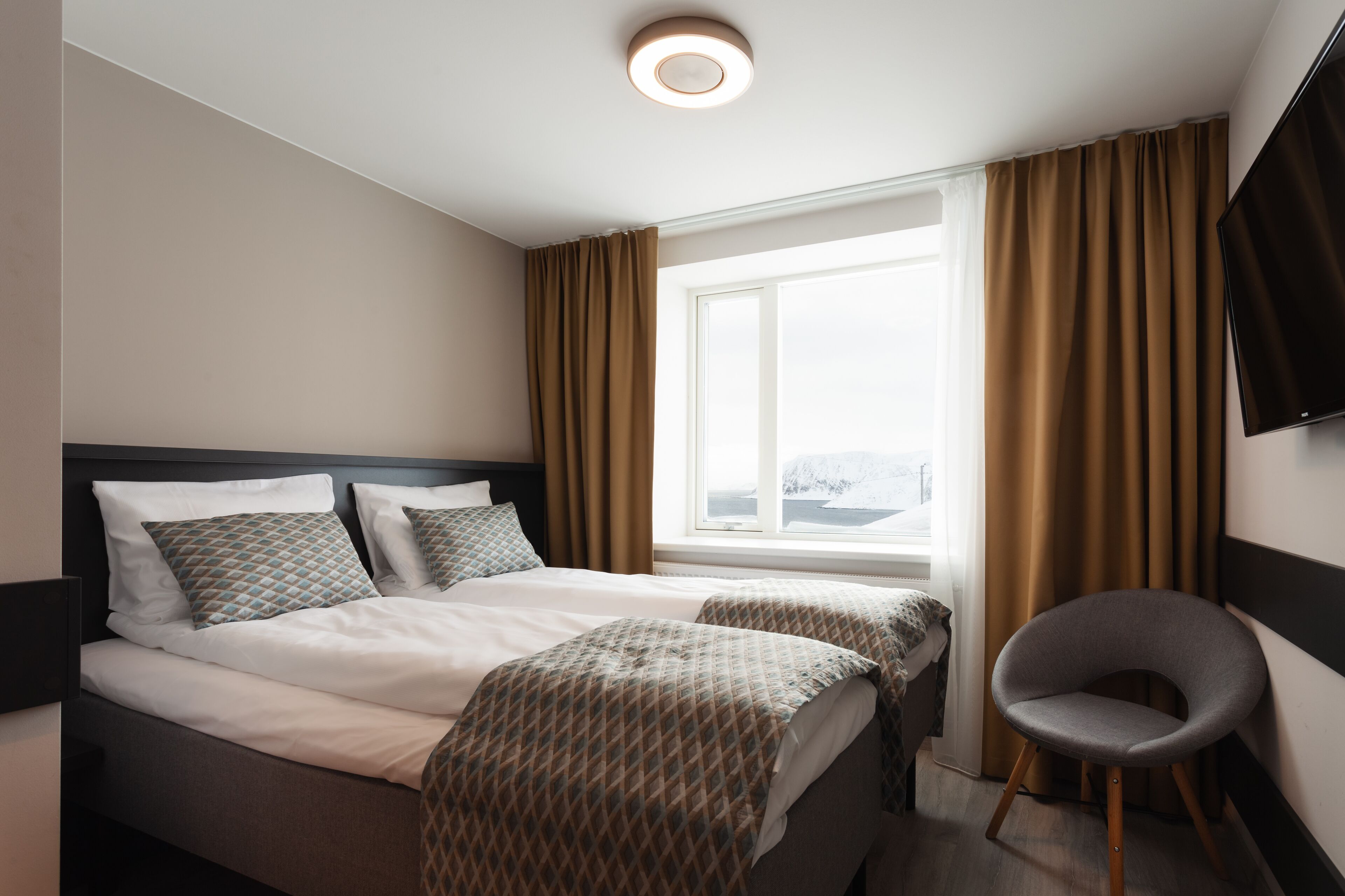 Ein Hotelzimmer mit zwei Betten, einem Fernseher, einem Sessel und einem Fenster mit Vorhängen.