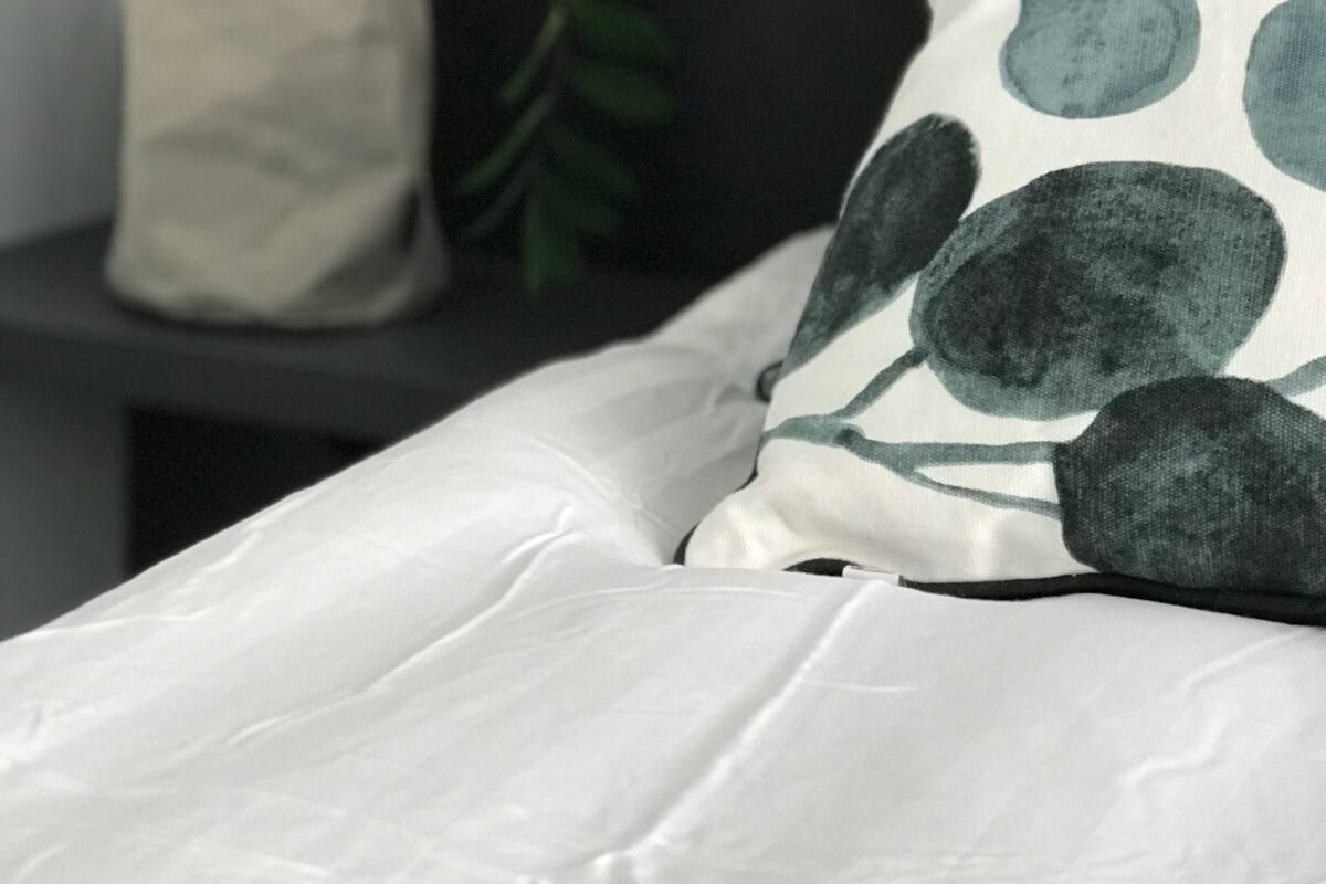 A bed with a white sheet and a pillow with a leaf pattern. There is a potted plant on a shelf beside the bed.