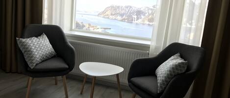 Ein Zimmer mit zwei schwarzen Sesseln mit gemusterten Kissen, einem kleinen weißen Tisch und Blick auf eine Küstenlandschaft durch ein großes Fenster.