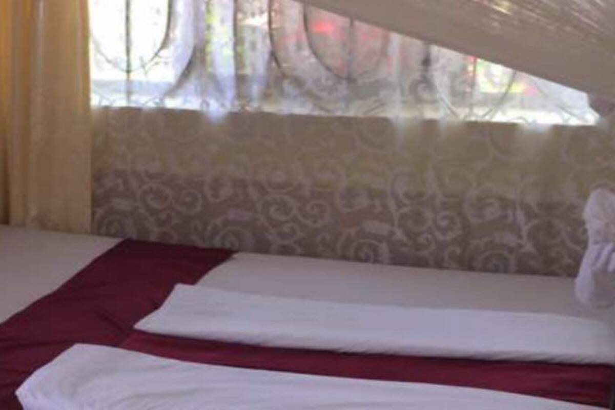 A single bed with white linens and a red accent, a headboard, and a window with patterned curtains.