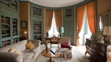 Ein Zimmer mit einer Couch, einem Sessel und einem Holzschrank. Ein Tisch mit Lampe und einem dekorativen Gegenstand steht dort. Das Zimmer hat ein Fenster mit Vorhängen und eine Bücherwand voller Bücher.