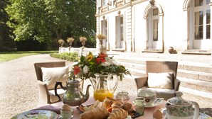 Una mesa de comedor al aire libre con una variedad de frutas, pasteles y una vajilla de té, con un edificio y árboles de fondo.