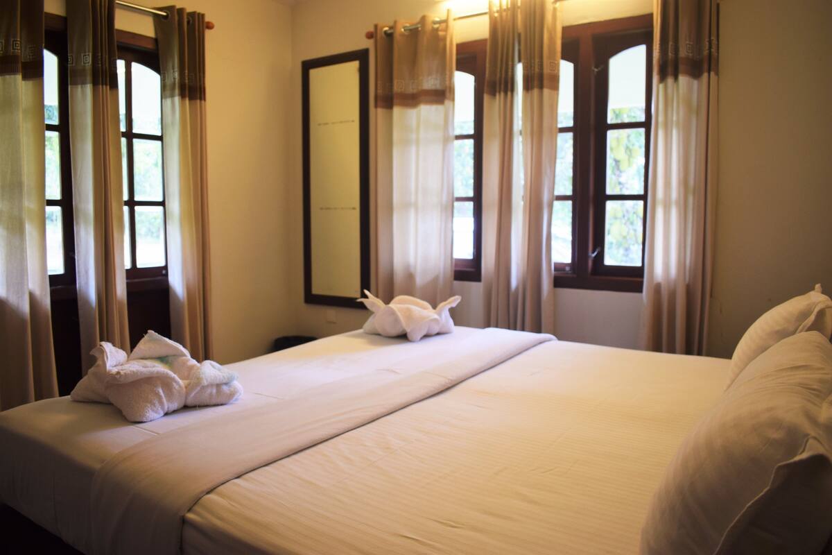 Habitación de hotel con una cama grande, dos ventanas con cortinas, un espejo y una toalla sobre la cama.