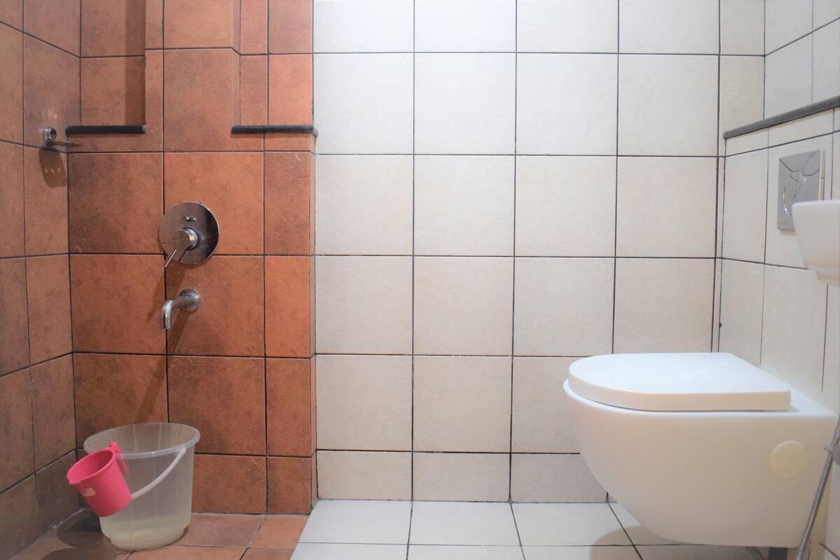 Un baño con inodoro blanco, zona de ducha con pared de azulejos marrones y un cubo de plástico transparente en el suelo.