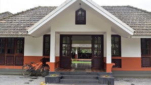 A building with a triangular roof, a central entrance with glass doors, and a bicycle parked outside. - 3 Hills County (Vythiri)