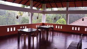 A covered dining area with a table and chairs, large windows, and a view of greenery outside. - 3 Hills County (Vythiri)