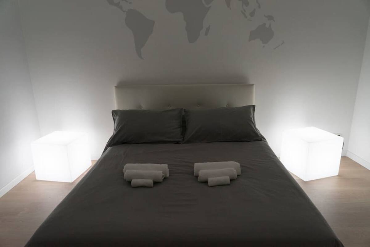 A neatly made bed with two pillows and two rolled towels, a world map on the wall, and two bedside tables with lamps.