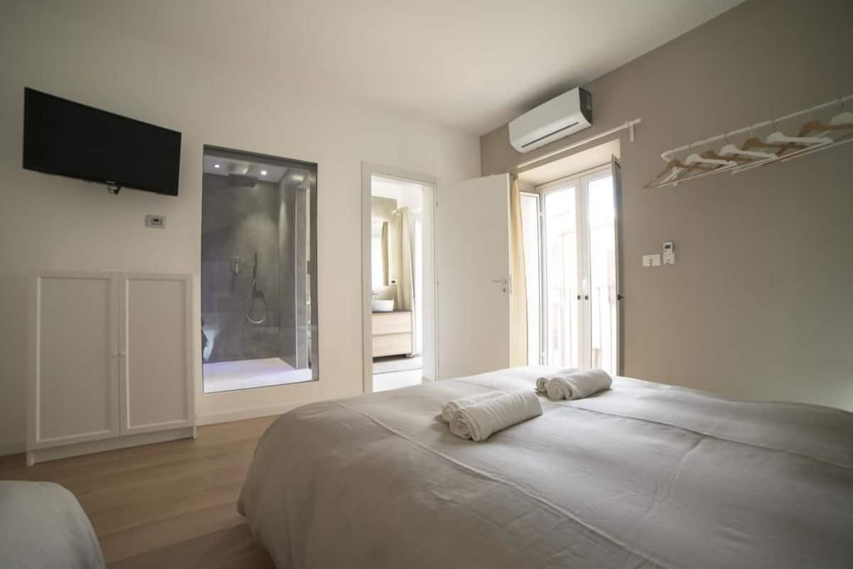 A bedroom with a large bed, a TV mounted on the wall, and a window overlooking a bathroom.