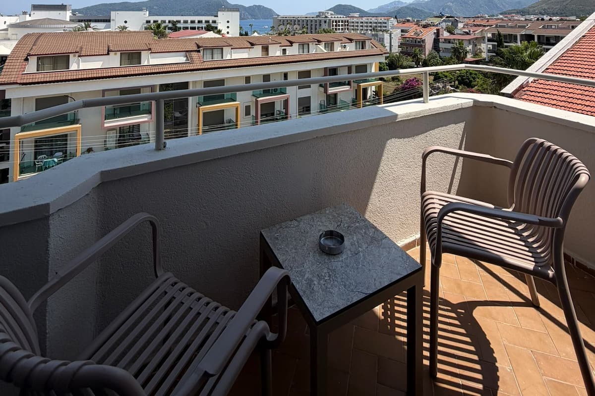 A balcony with a table and chairs, overlooking a cityscape with buildings and mountains.