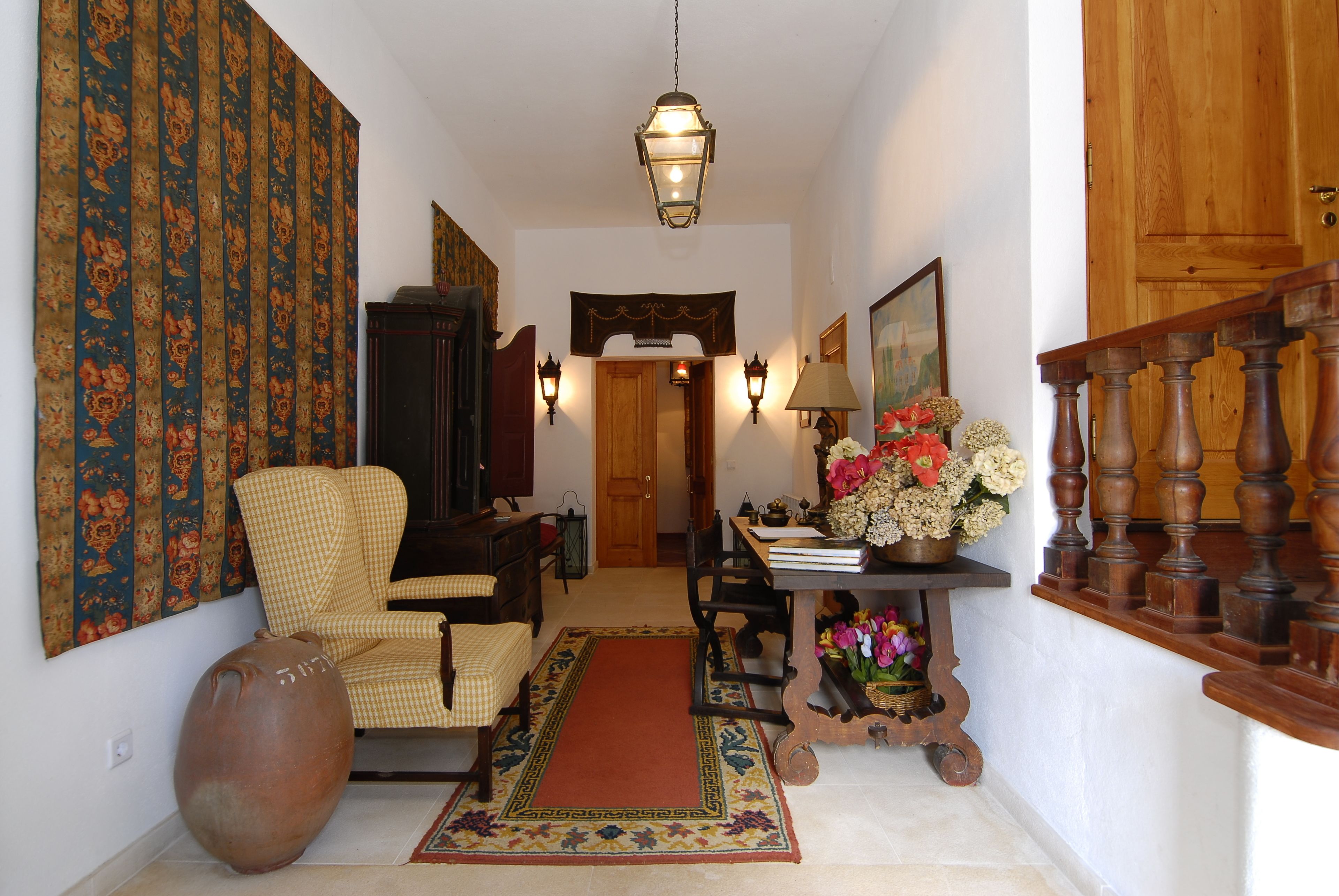 A hallway with a checkered armchair, a large vase, a wooden table with flowers, and a staircase with wooden balusters.